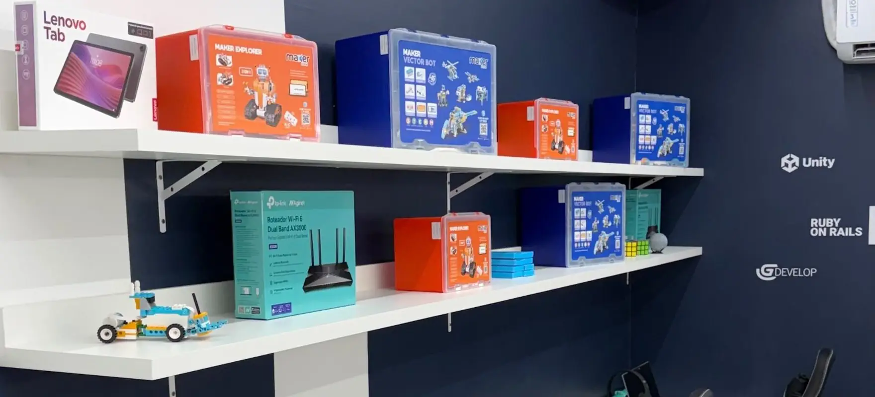 Sala de aula da unidade de Joinville com kits de robótica na prateleira e estações com notebook